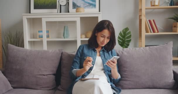 Hermosa chica escribiendo en un cuaderno mirando la pantalla del teléfono inteligente sentado en casa — Vídeo de stock