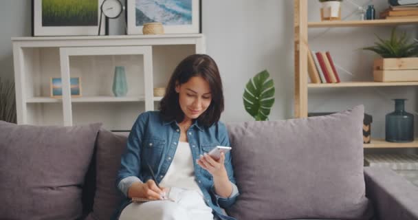 Mujer joven sosteniendo el teléfono inteligente mirando la pantalla y escribiendo en un cuaderno — Vídeo de stock