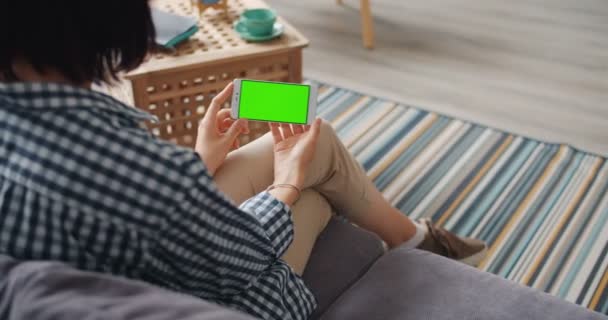 Jeune femme utilisant un smartphone avec écran vert maquette assis sur le canapé à la maison — Video