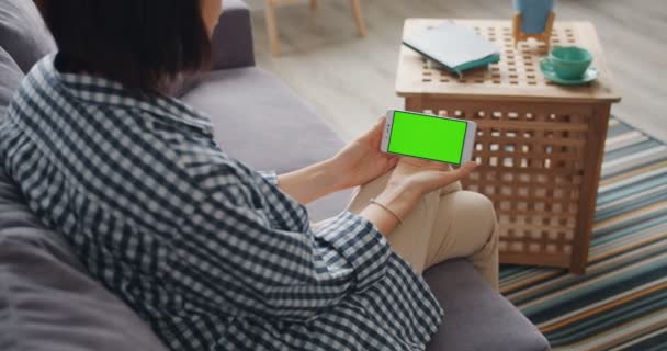 Chica mirando la pantalla de maqueta verde del teléfono inteligente sentado en el sofá en casa — Vídeo de stock