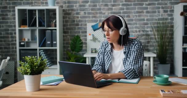 Симпатичная девушка слушает музыку в наушниках, танцуя с ноутбуком в офисе — стоковое видео