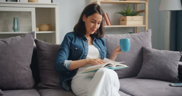 Милая студентка читает книги и пьет кофе расслабляясь дома на диване — стоковое видео