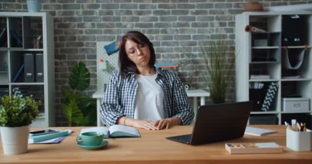 Уставшая девушка работает с ноутбуком в офисе, а затем растягивает руки расслабляясь на работе — стоковое видео