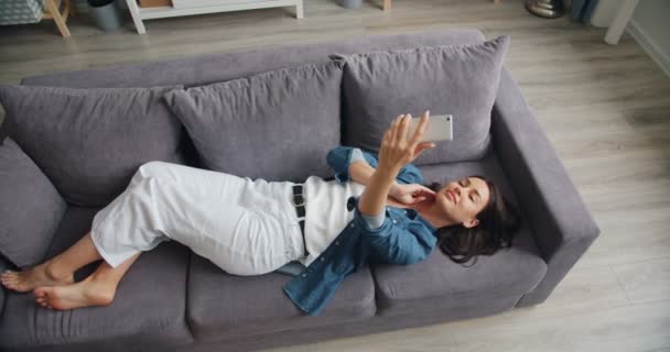 Pohled na krásnou ženu, která si bere s kamerou smartphone a leží na gauči — Stock video