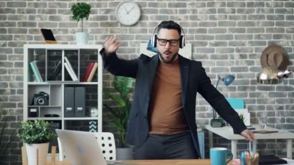 Funny businessman relaxing at work listening to music in headphones dancing — Stock Video