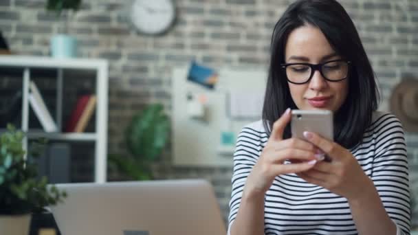 Привлекательная юная леди касается экрана смартфона улыбаясь на рабочем месте — стоковое видео