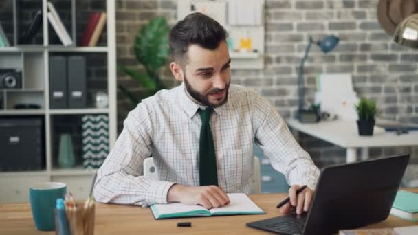 Tipo barbudo usando la escritura del ordenador portátil en el cuaderno sentado en la mesa en la oficina — Vídeo de stock