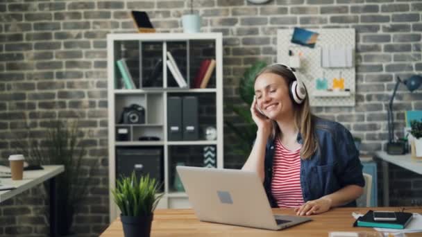 Pretty lady in headphones listening to music in office dancing raising arms — Stock Video