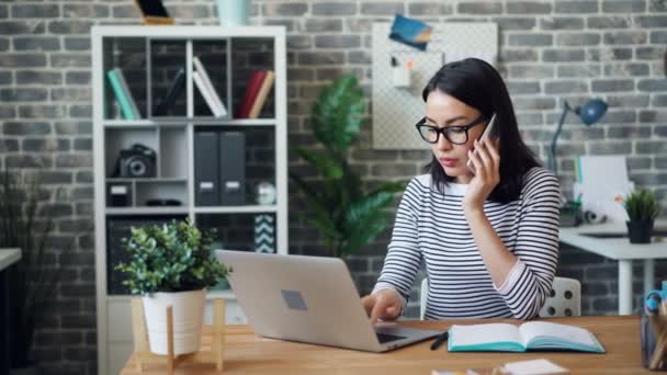 Vrouwelijke kantoormedewerker praten op mobiele telefoon en het gebruik van laptop dan het nemen van notities — Stockvideo
