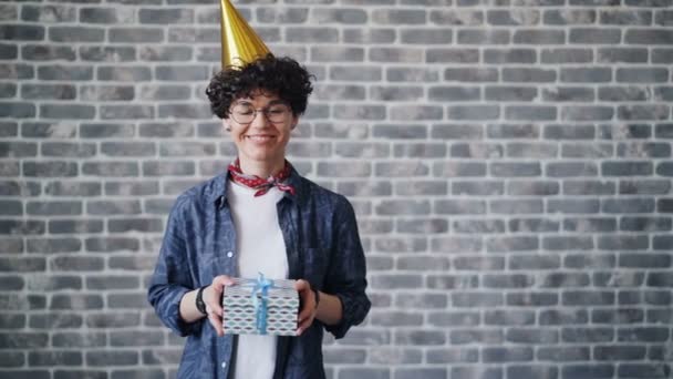 Портрет веселої молодої жінки в яскравому вечірньому капелюсі, що тримає подарункову коробку посміхаючись — стокове відео