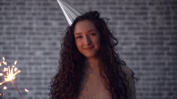 Donna sorridente in cappello partito tenendo luce bengala ballare su sfondo di mattoni — Video Stock