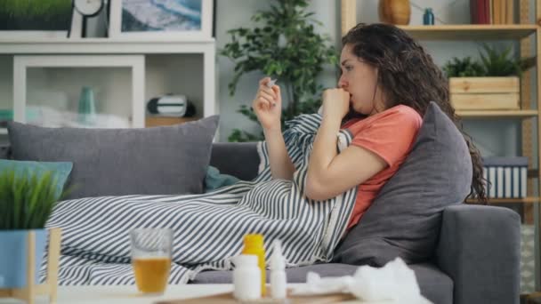 Sidovy av ung kvinna tar temperatur med termometer i huset på soffan — Stockvideo