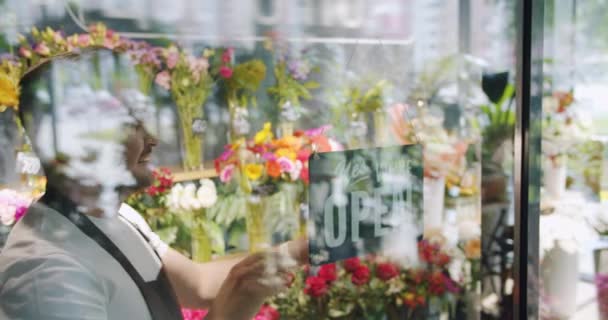 Chico guapo en delantal cambiante abierto para cerrar letrero en ventana de la tienda de flores — Vídeo de stock