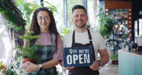Portrait of girl and guy holding green plant and open sign in flower store — Stock Video