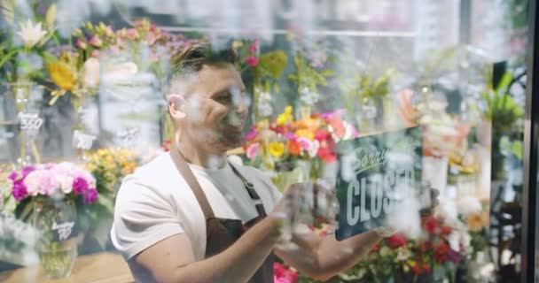 Florista masculino em avental mudando sinal fechado para abrir na porta da loja de flores sorrindo — Vídeo de Stock