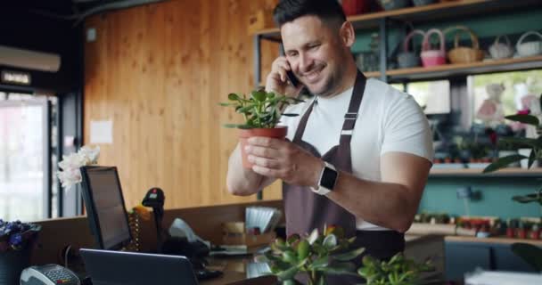 Floristería masculina hablando por teléfono móvil sosteniendo planta verde discutiendo orden — Vídeo de stock