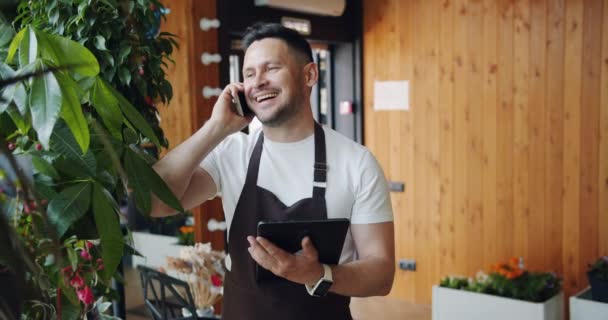 Leende Guy florist talar på mobiltelefon och håller modern Tablet på jobbet — Stockvideo