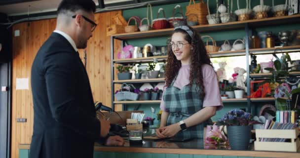 Радостный парень платит за цветы в магазине, используя кредитную карту, разговаривая с флористом — стоковое видео