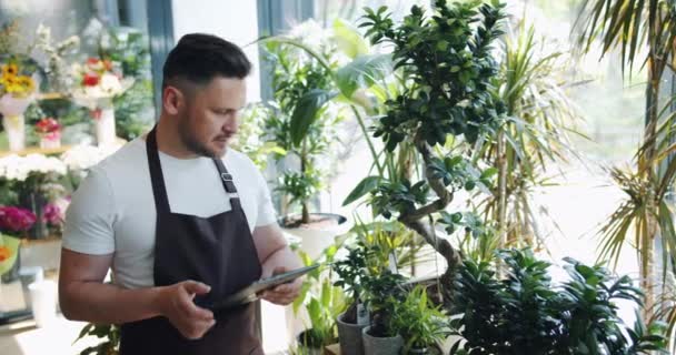 Feliz florista masculino trabajando con la tableta en la tienda de flores sonriendo mirando a las plantas — Vídeo de stock