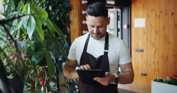 Hombre guapo en delantal disfrutando de flores en floristerías usando tabletas — Vídeos de Stock