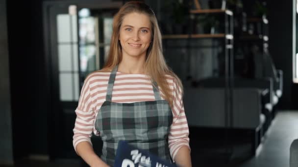 Retrato de chica bonita de pie en la cafetería sosteniendo letrero abierto dando la bienvenida a los clientes — Vídeo de stock