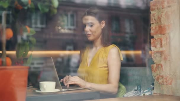 Bella dama usando el ordenador portátil en la cafetería escribiendo sonriendo en la mesa detrás de la ventana de cristal — Vídeo de stock