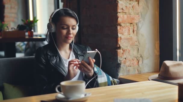 Happy female student listening to music in headphones using smartphone in cafe — Stock Video