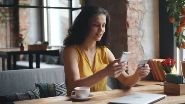 カフェに一人で座っているスマートフォンやクレジットカードでオンラインで支払う興奮した女の子 — ストック動画