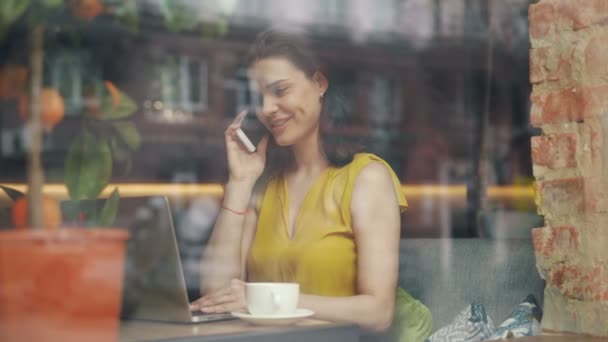 Привлекательный студент разговаривает по мобильному телефону, работает с ноутбуком в современном кафе — стоковое видео