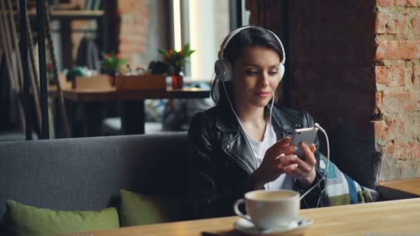 Mujer joven con auriculares escuchando música usando smartphone en la cafetería moderna — Vídeo de stock