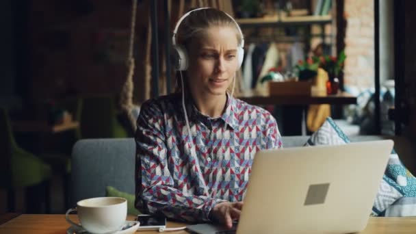 Радостная девушка слушает музыку в наушниках, используя ноутбук, печатая песни в кафе — стоковое видео