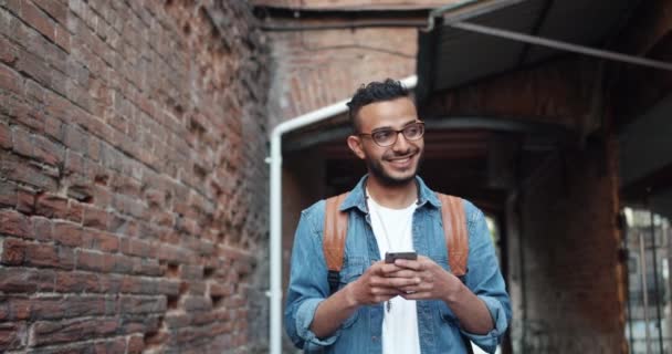 ハンサムなひげアラブは、屋外で笑顔の現代のスマートフォンアプリケーションを楽しんでいます — ストック動画