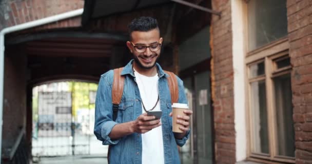 Alegre joven árabe tocando la pantalla del teléfono inteligente sosteniendo la taza de café al aire libre — Vídeo de stock