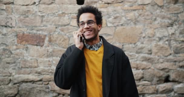 Mouvement lent de l'homme afro-américain joyeux parlant sur un téléphone mobile à l'extérieur — Video
