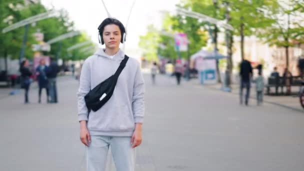 Zoom-out time-lapse de chico lindo en los auriculares de pie en la calle en la ciudad — Vídeos de Stock