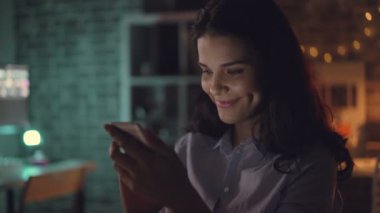 Neşeli genç kadın karanlık ofiste geceleri gülümseyerek akıllı telefon kullanarak