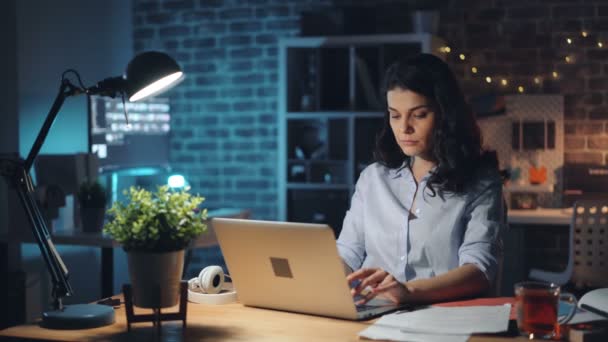 Молода жінка вимикає ноутбук і світло в офісі вночі і залишає роботу — стокове відео