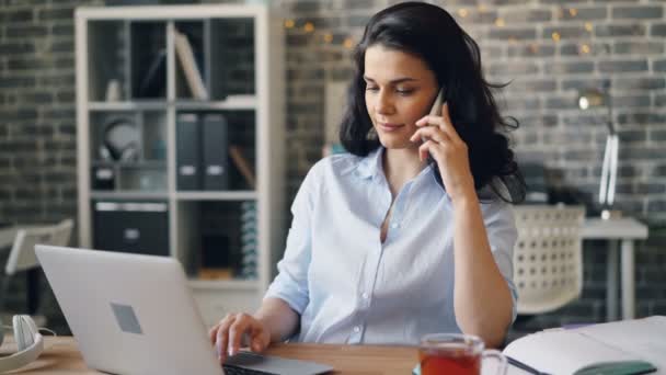 Mooi meisje spreken op mobiele telefoon met behulp van laptop en glimlachend in Office — Stockvideo