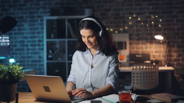 Menina bonito trabalhar com laptop e desfrutar de música no fone de ouvido no escritório à noite — Vídeo de Stock