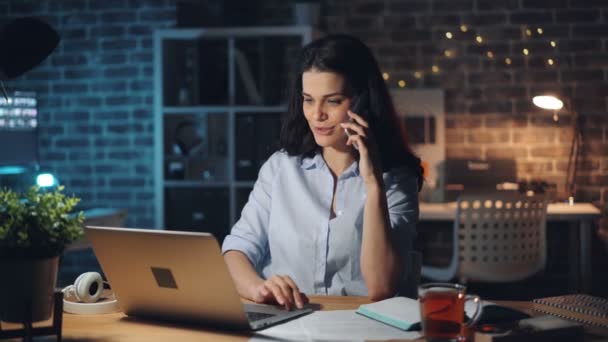 Mooie kantoormedewerker chatten op mobiele telefoon in Office met behulp van laptop 's nachts — Stockvideo