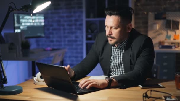 Erfolgreicher Geschäftsmann arbeitet nachts mit Laptop im Büro und lächelt entspannt — Stockvideo