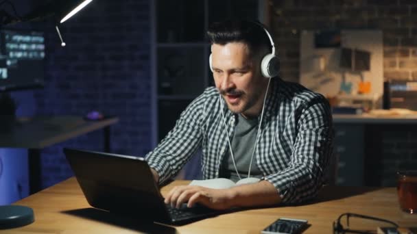 Trabajador de oficina feliz en auriculares escuchando música ocupada con el ordenador portátil por la noche — Vídeos de Stock