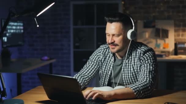 Улыбающийся парень слушает музыку с наушниками, используя ноутбук в офисе ночью — стоковое видео
