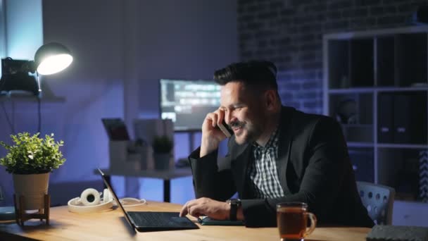 Wesoły pracownik biurowy rozmawiający na telefon i pracujący z laptopem w ciemnym biurze — Wideo stockowe