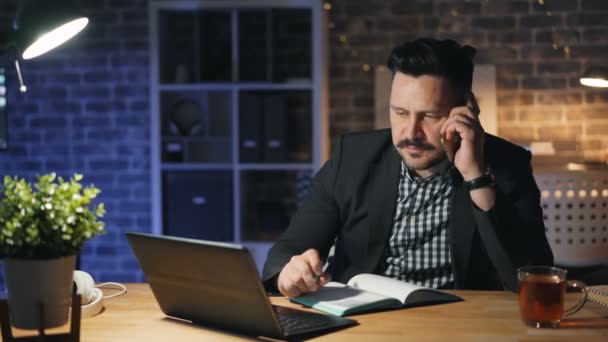 Homme sérieux employé de bureau parlant sur un téléphone mobile et utilisant un ordinateur portable la nuit — Video