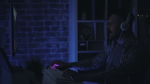 Aufgeregter Kerl mit Kopfhörern spielt Computervideospiel zu Hause spät in der Nacht — Stockvideo
