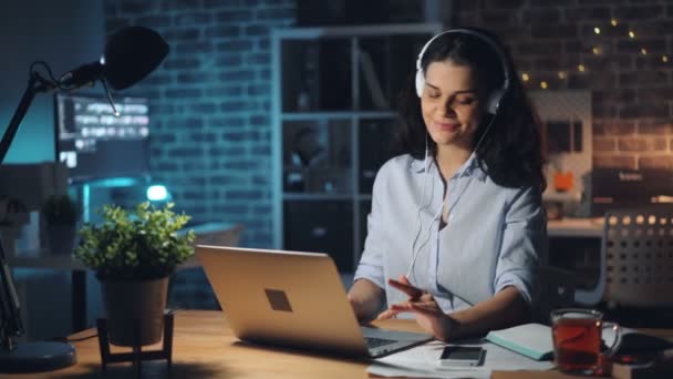 Bonita senhora em fones de ouvido ouvindo música no escritório escuro trabalhando com laptop — Vídeo de Stock