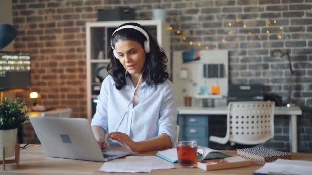 Chica feliz trabajando con el ordenador portátil escribiendo y escuchando música a través de auriculares — Vídeos de Stock