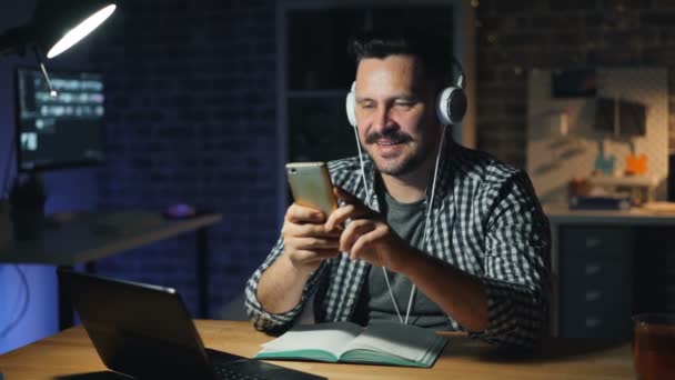 Tipo atractivo usando el teléfono inteligente y escuchar música en la oficina por la noche — Vídeos de Stock