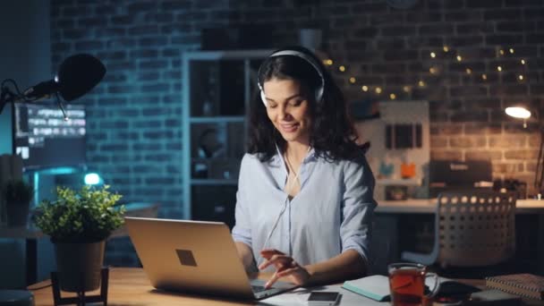 Mulher digitando no laptop ouvindo música em fones de ouvido no escritório escuro à noite — Vídeo de Stock